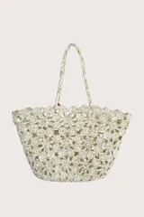 Bolso tipo cesta tejido a crochet con diseño de flores caladas en color blanco y detalles dorados. Cuenta con asas trenzadas y una etiqueta decorativa de cuero sintético marrón.