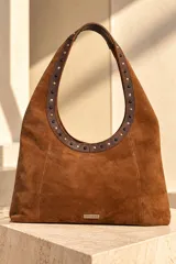 Cartera tipo hobo de gamuza color marrón chocolate, con asa de hombro decorada con tachas metálicas en tonos dorados, plateados y negros, y placa con el logo de la marca en la parte inferior frontal.