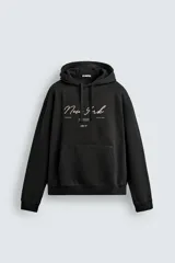 Sudadera de corte relajado color negro con capucha ajustable y bolsillo frontal tipo canguro. Presenta estampado gráfico en el frente con la inscripción New York y detalles adicionales en contraste.