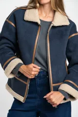 Campera tipo gamulán de corte oversize, color azul marino con interior y cuello de corderito en tono beige. Presenta detalles de costuras y bordes en cuero sintético color marrón, cierre frontal y mangas largas.