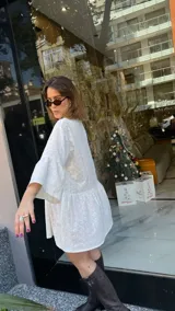 Vestido corto de broderie color blanco, con cuello camisero, cierre frontal de botones y mangas tres cuartos. Presenta un diseño de corte holgado con falda de volados y ruedo irregular.