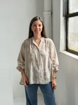 Camisa de lino color beige con diseño oversize, cuello camisero mao, cierre frontal con botones ocultos, mangas largas abullonadas con puños elásticos y delicados bordados florales tono sobre tono en el frente y las mangas.
