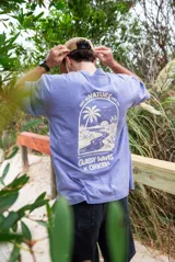 Remeron de corte oversize color violeta lavado, con estampa gráfica en la espalda que representa un paisaje natural con palmeras y un río, junto con el texto 'Part of Nature like you' y 'Glassy Waves x Origen'.