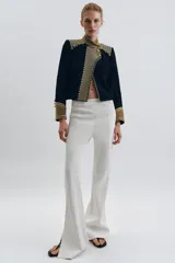 Chaqueta de estilo militar en color negro, con cuello subido y puños en terciopelo color verde oliva. Presenta detalles de alamares dorados en el frente y hombros, bolsillos con solapa y cierre frontal con botones metálicos en relieve.