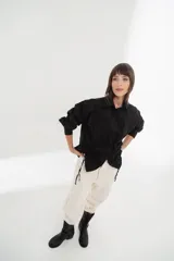 Camisa blanca de mangas largas con cuello clásico y cierre con botones. Presenta detalles de fruncido vertical en la parte delantera y cordones ajustables en los laterales. Corte holgado.