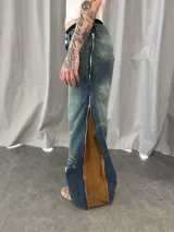 Jean de corte amplio con lavado desgastado en tono azul verdoso. Presenta bordados lineales en ambas piernas y cierres laterales metálicos que, al abrirse, revelan un panel de pana en color marrón. Cuenta con bolsillos amplios y botones personalizados.