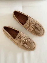 Mocasín tipo náutico de gamuza color camel, con detalle de flecos y borlas en el empeine, cordones laterales y suela plana de goma.