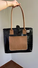 Bolso tote negro con textura símil cuero de cocodrilo, bolsillo externo color beige y asas marrones.