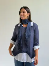 Sweater de punto azul con diseño asimétrico y detalles bordados, que incluye una bufanda a juego y una camisa blanca de lino debajo.