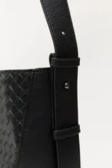 Cartera de hombro color negro con diseño trenzado en el panel frontal y laterales lisos. Cuenta con una correa larga ajustable y una placa metálica pequeña con el logo de la marca en la parte inferior.