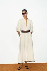 Vestido midi color crudo, con cuello camisero, mangas abullonadas por debajo del codo con puños elásticos y cierre frontal oculto con botones. Incluye bolsillos laterales y un cinturón ancho de cuero marrón oscuro con hebilla metálica.