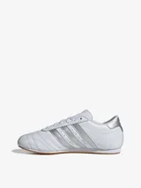 Zapatillas Adidas Taekwondo Lace blancas con detalles plateados en las tres tiras laterales y el talón. Diseño de perfil bajo con puntera pespunteada y suela de goma color caramelo.