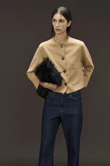 Chaqueta corta color camel de gamuza sintética, con calce holgado, cuello redondo, acceso por medio de botones en cartera central, bolsillos plaqué en delantero y manga larga con recortes.