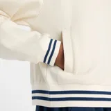 Campera deportiva New Balance color blanco hueso, con cierre frontal, cuello alto y puños y cintura con ribetes a rayas azul marino. Presenta un pequeño logo bordado en el pecho.