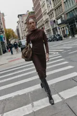 Vestido midi de manga larga, color marrón oscuro, confeccionado en tejido de encaje o punto con patrón floral sutil. Presenta cuello perkins y drapeado asimétrico en el torso con frunces laterales.