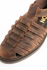 Sandalias cangrejeras de cuero color marrón, con diseño de tiras cruzadas y cierre de hebilla metálica en el lateral. Poseen suela plana y gruesa.