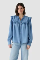Blusa de manga larga en tejido denim celeste, con cuello mao y volados fruncidos en los hombros y a lo largo de la botonera frontal. Presenta puños ajustados con elástico.
