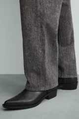 Bota de media caña en color negro, con diseño de arnés que incluye tiras, argollas metálicas y tachas decorativas alrededor del tobillo. Presenta punta fina y tacón cuadrado.