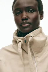 Campera beige en mezcla de lino, con cuello elevado con cordón ajustable, cierre frontal y bolsillos laterales con solapa y botón. Presenta hombros caídos y elástico revestido en el bajo.