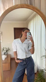 Blusa celeste a rayas blancas de lino, con cuello marinero amplio con bordes ondulados, mangas cortas abullonadas y doble botonadura frontal con botones blancos.