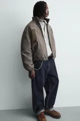 Cazadora de corte boxy fit confeccionada en tejido técnico. Presenta un diseño a rayas verticales, cuello subido, cierre frontal de cremallera y mangas largas con puños elásticos. Incluye bolsillos laterales con cremallera oculta y detalles de paneles en contraste.