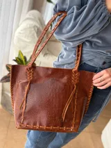 Cartera tipo shopper de cuero genuino color marrón con estampado texturado tipo cocodrilo. Presenta detalles de costuras y tiras trenzadas artesanales en los bordes y en las asas de hombro.