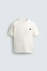 Camiseta de corte relajado (relaxed fit) color blanco hueso, con cuello redondo, manga corta y bolsillo plastrón en el pecho. Presenta un bordado pequeño y contrastante de una palmera en el área del bolsillo.