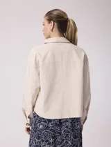 Chaqueta de lino color beige, de corte recto y holgado, con mangas largas, cuello camisero y cierre frontal de botones carey. Cuenta con bolsillos en el pecho y puños abotonados.