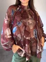 Camisa de seda italiana con estampado floral, cuello alto, mangas largas abullonadas y volados frontales. Presenta un lazo para atar en la espalda.