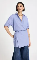 Vestido camisero corto de algodón en color celeste con finas rayas blancas, con diseño asimétrico tipo wrap, mangas largas y escote en V con cuello camisero.