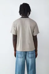 Camiseta de manga corta con corte amplio y diseño de costuras decorativas tipo piping en el frente. Presenta cuello redondo en color negro en contraste con el cuerpo de la prenda en tono beige.