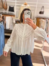 Blusa blanca de algodón con textura rayada, cuello alto con volado y cierre frontal de botones. Presenta detalles de puntilla calada en el pecho y mangas largas abullonadas con puños ajustados.