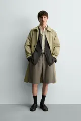 Trench de corte relajado confeccionado en tejido técnico italiano, color beige. Presenta cuello camisero, manga larga, bolsillos de vivo en la cadera y cierre frontal de botonadura simple con botones oscuros.