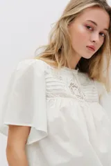 Blusa blanca de algodón con cuello redondo ribeteado en encaje y mangas cortas holgadas. Presenta un canesú fruncido con jaretas y detalles de broderie en la parte frontal.