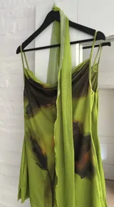 Vestido corto de tirantes finos con escote drapeado, confeccionado en tela semitransparente con estampado abstracto en tonos verdes y negros. Incluye un accesorio tipo pañuelo integrado al cuello.