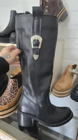 Bota alta de cuero marrón, con diseño de inspiración ecuestre, taco ancho y hebilla metálica decorativa en la parte superior de la caña.