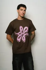 Remera de manga corta, corte regular fit, color marrón oscuro con un gran estampado frontal en rosa pálido que simula un logo estilizado o un símbolo abstracto.