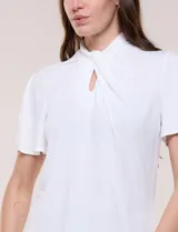 Blusa blanca de corte recto con cuello alto torzado, abertura frontal en gota y mangas cortas con aberturas sobre los hombros.