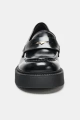 Mocasín de cuero negro con suela gruesa tipo plataforma, detalle de antifaz con aplique metálico dorado y costuras reforzadas en la puntera.