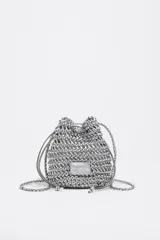 Bolso mini tipo saco confeccionado con tiras entrelazadas color plateado, con cierre de cordón ajustable y correa larga trenzada.
