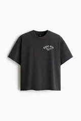 Remera boxy de algodón gris oscuro, con corte oversized, hombros caídos, cuello redondo ribeteado y estampa pequeña en el pecho con texto en blanco.
