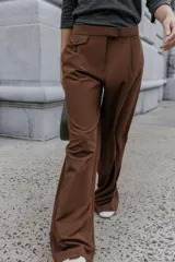 Pantalón de vestir marrón oscuro, tiro alto, con pernera ancha y pinzas frontales. Presenta un bolsillo con tapeta en la parte superior delantera.