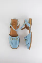 Sandalias de cuero color celeste, con punta cerrada, flecos frontales y detalle de estribo metálico dorado. Tienen tira ajustable al tobillo y suela plana con pequeño taco.