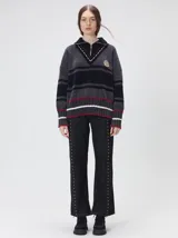 Pullover de punto con textura aterciopelada, diseño oversize, cuello polo con cierre frontal parcial y detalles de pespuntes en contraste. Presenta rayas horizontales en tonos tierra y un parche bordado con la letra M en el pecho.