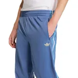 Pantalón deportivo Adidas Firebird de tejido liso, color azul claro, con cintura elástica y tres franjas laterales en tonos de azul y blanco. Presenta un pequeño logo de trébol dorado bordado en el muslo izquierdo y un bolsillo lateral con cierre.