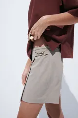 Pollera mini de corte cruzado con diseño a cuadros en tonos beige y marrón. Presenta una hebilla metálica decorativa en el lateral y cierre oculto en la parte posterior.
