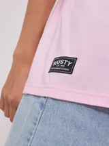 Remera de corte oversize color rosado, con cuello redondo y mangas cortas. Presenta un estampado frontal con la frase 'surf girl' y el logo de la marca Rusty, acompañado de un diseño floral.