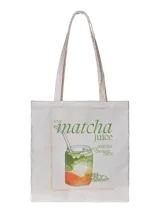 Bolso tipo tote bag de color blanco crudo, confeccionado en mezcla de algodón y poliéster. Presenta un diseño gráfico frontal con ilustración de un vaso de matcha con naranja y tipografía en tonos verdes. Cuenta con dos asas largas para llevar al hombro.