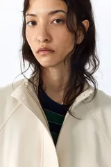 Campera corta de lino color marfil con cuello alto, solapa con botones, bolsillos diagonales con solapa, mangas raglán y cordón ajustable en la cintura.