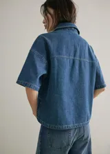 Blusa de algodón estilo denim, con diseño oversize, cuello camisero, mangas cortas, dos bolsillos de parche en el pecho y cierre frontal de botones.